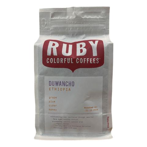 A white bag of coffee with a red logo up top that says the brand, "Ruby Colorful Coffees". There is a label on the bag with the name of the beans - Duwancho - and it says Ethiopia underneath. It lists flavor notes - grape, plum, cider, and honey - as well as additional inromation about the relationship, farm type, processing, region, variety, and elevation.