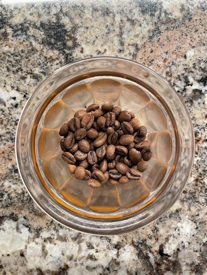 An overhead shot of a small glass containing little coffee beans. The glass sits on a marble countertop.