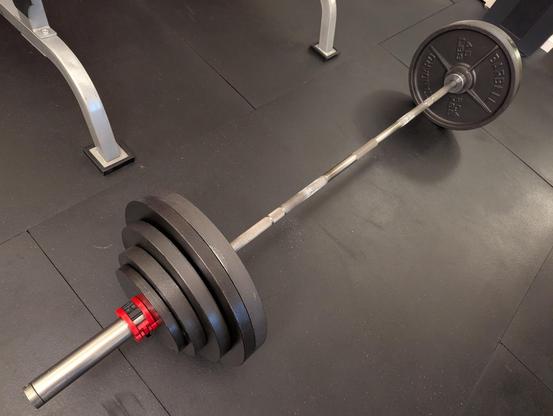 Photograph of a loaded barbell on a black rubber mat floor.
Each side of the barbell has two 45, one 35, one 25, one 10 and one 5 lb plate held in place with a red collar.
In the top right corner is a black HEPA air filter.