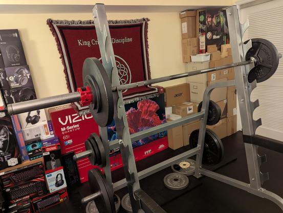 Photograph of a loaded barbell on a sawtooth combination rack on a black rubber mat floor.
The bar is racked at shoulder height.
Each side of the barbell has one 45, one 10 and one 5 lb plate held in place with a red collar.
Behind the rack are a collection of many electronics product boxes against a pale yellow wall providing essential acoustic treatment and a hanging King Crimson "Discipline" rug.