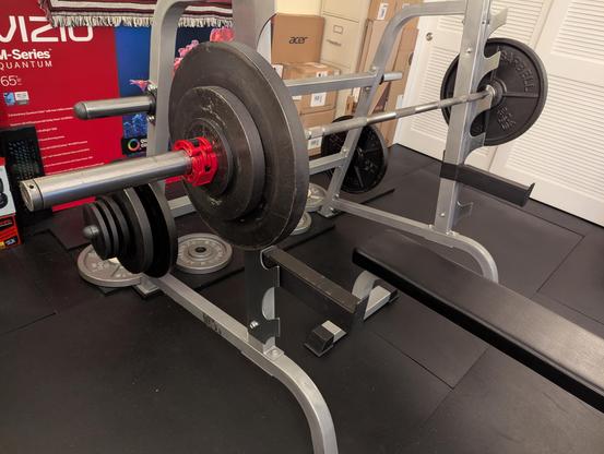 Photograph of a loaded barbell on a sawtooth combination rack on a black rubber mat floor.
The bar is racked at waist height.
Under the bar is a lifting bench with black padding.
Each side of the barbell has one 45, one 25, and one 2.5 lb plate held in place with a red collar.
Behind the rack are a collection of many electronics product boxes against a pale yellow wall providing essential acoustic treatment and a hanging King Crimson "Discipline" rug.