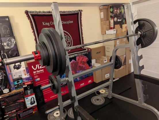 Photograph of a loaded barbell on a sawtooth combination rack on a black rubber mat floor.
The bar is racked at shoulder height.
Each side of the barbell has two 45, one 35, one 10 and two 5 lb plates held in place with a red collar.
Behind the rack are a collection of many electronics product boxes against a pale yellow wall providing essential acoustic treatment and a hanging King Crimson "Discipline" rug.