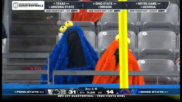 Two fans dressed in large, colorful monster costumes, one blue with yellow eyes and the other orange, are sitting in a stadium. They are watching a college football game, with scoreboard details visible in the foreground.