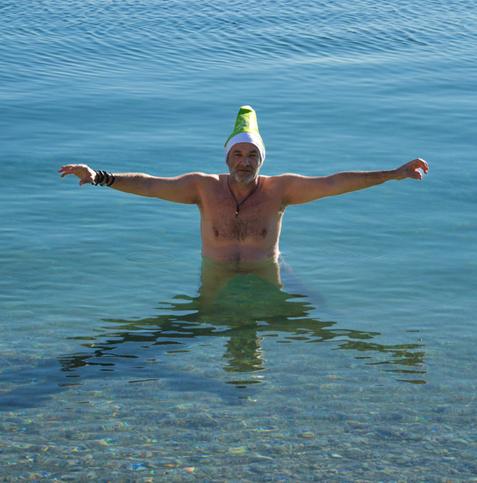 Mann mit grüner Weihnachtsmütze badet im Meer und streckt die Arme aus