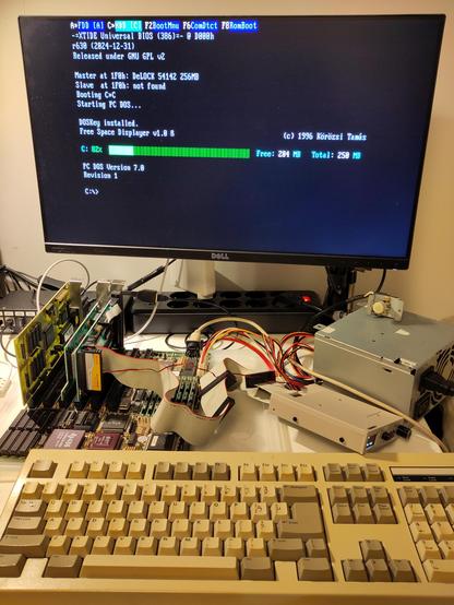 The 486DLC motherboard laid on the desk, with 3 cards inserted, a VGA, an Ethernet and a multi I/O card. The system is booted into PC-DOS 2000 (a.k.a. 7.0 Revision 1). The screen shows the boot messages, but also the XT IDE ROM, seated on the Ethernet card, detecting the DoM hooked on the IDE controller.