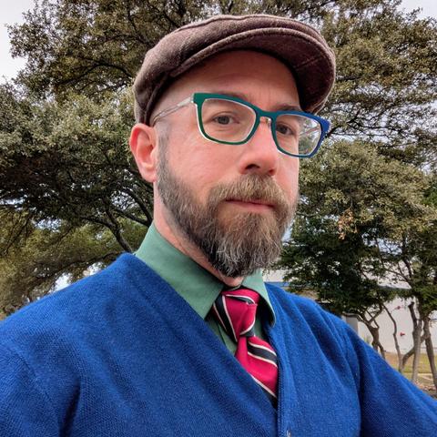 A photo of me, a 46 year old white man with a shaved head, a short cropped brown & grey beard, 

photographed from the chest up in front of some live oak trees in an office park,

wearing

a rectangle glasses with blue & green frames, a green dress shirt, a deep red necktie with dark blue diagonal bands in a trinity knot, under a navy blue cardigan and a tan wool flat cap.
