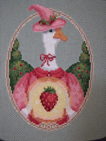 A completed cross stitch showing a goose in a pink witch hat with a bow around her neck. She is wearing a pink frock and holding a glowing strawberry. She has a stitched frame around her and some strawberry bushes in the background.
