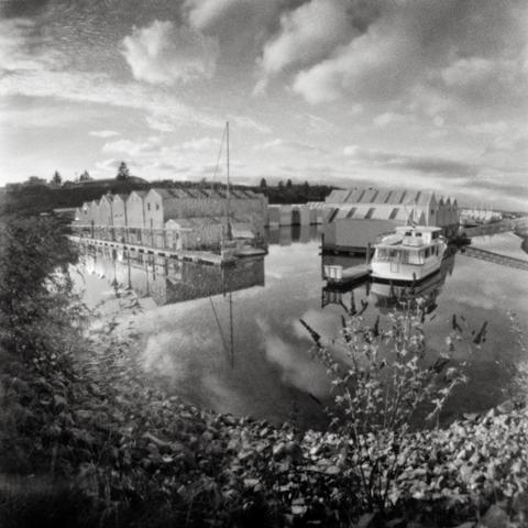 Monochrome pinhole image of boat houses on calm water with cloud reflections.