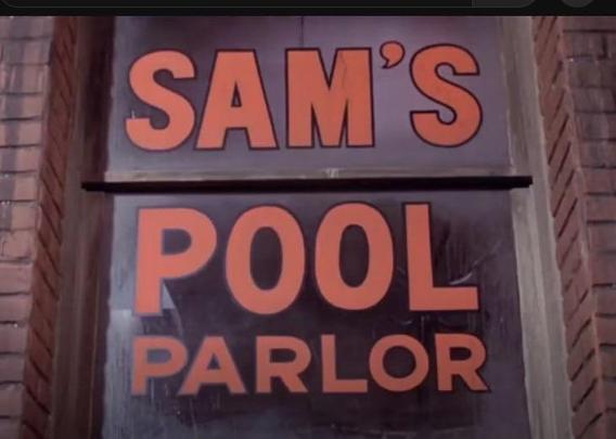 Sign reading "Sam's Pool Parlor" in bold, orange letters on a window against a brick wall.