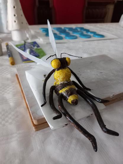 Foto a una abeja hecha en pasta de modelar y alambre y pintada con acrílicos amarillos y marrón oscuro. La abeja está sobre papel de horno y una tablilla de madera encima de una mesa. Al fondo se ven difuminados círculos azules en pasta de modelar.