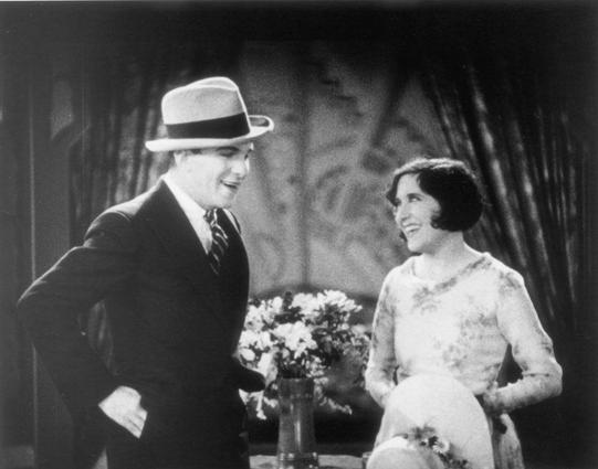 A black-and-white still of husband and wife comedy team George Burns and Gracie Allen in the 1929 transitional sound comedic short “Lambchops”. Burns is waring a dark suit and a hat and Allen is waring a floral, tea-length dress with modest high heels. They are talking and making each other smile.