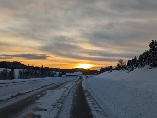 Die Sonne geht gerade unter, der Himmel ist leicht bewölkt und zeigt sich in verschiedenen Gelb- und Orangetönen. Straße und Landschaft sind voll Schnee.