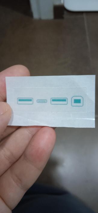 A white left hand holds a small, white sticker with four teal-colored symbols. The symbols appear to represent different types of ports or conectors:
USB A - USB C - USB A - USB B
The sticker uses geek symbols to represent the acronym ACAB