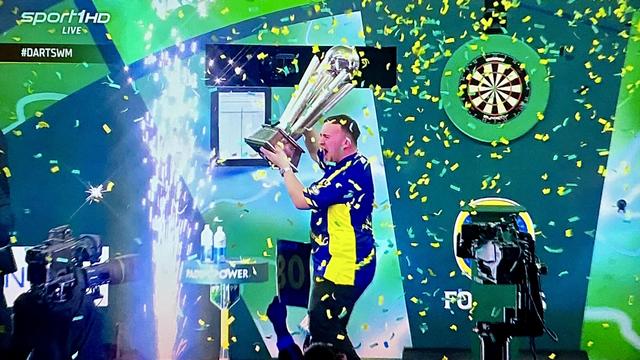 Luke Littler jubelt mit WM-Pokal auf der Bühne, Konfetti fliegt auch rum.