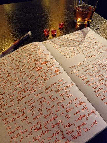 An open notebook with writing in red ink, dice and a pint glass with a bit of cider in it on a high bar table