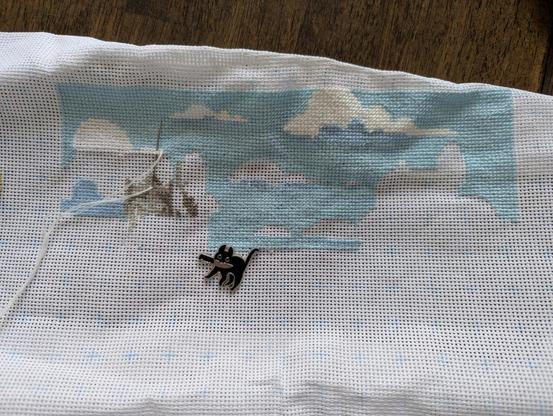 Am in-progress cross stitch project. White Aida fabric with about 4,000 light blue stitches for the sky, with some white clouds. A needle minder in the shape of a black cat holding a knife in its mouth