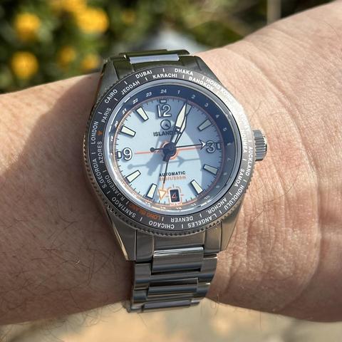 Watch with brushed stainless steel case with polished chamfer, brushed stainless steel H-link bracelet, world time bezel, pale blue dial with white numeric indices outlined in black at 12, 3 and 9, orange-on-brown date at 6:00 with date window outlined in orange, baton indices elsewhere, skeletonized black sword hands with white lume, orange GMT hand, arrow seconds hand, and 24-hour markings on the rehaut. 