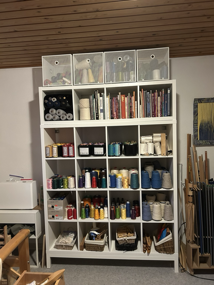 A white storage unit with 4x5 cubbies filled with weaving yarn, weaving books, and, in the lowest row, weaving tools. The top of the storage unit has 4 white metal baskets holding more weaving yarn