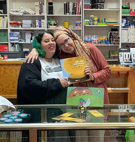 Foto hecha a Arancha Ferrero y a mí riendo abrazadas mientras sujetamos un ejemplar de "Una cosa que volaba" entre las 2 detrás de un mostrador con una de las ilustraciones del álbum ilustrado dedicada por Arancha, una abejita hecha en pasta de modelar, varios marcapáginas y unos colgandines hechos en pasta de modelar con cuerda de yute verde.