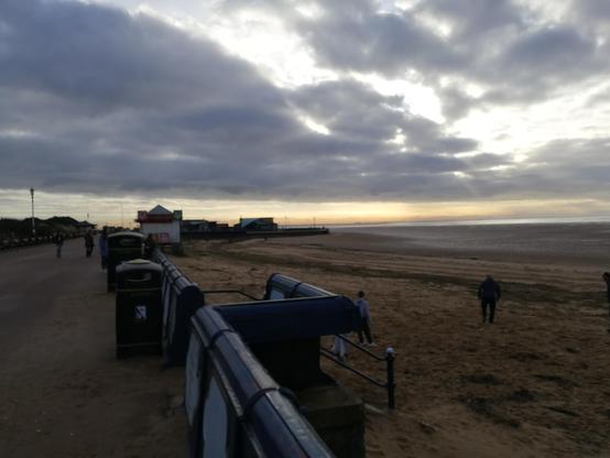 Early morning view of St Annes promenade and beach. The image shows a cloudy sky with the sun peeking through, casting a soft light over the beach. The promenade has a few people walking around. The beach is mostly empty, with a few people scattered around. In the distance, there are some buildings and structures near the shoreline.