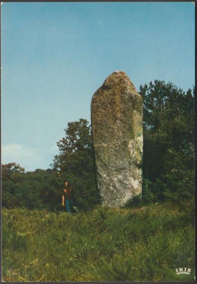 A relatively modern colour postcard showing the Menhir de Ceinturat, a 5.1m or 16¾ft tall prehistoric standing stone by Cieux near Limoges in France. A man stands beside the menhir and looks very small in comparison. The stone stands of a slight bank amidst tall grass and there is a line of trees behind the stone. The sky is blue and clear.
