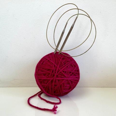 A vibrant dark pink ball of yarn, sitting on a white background, with a circular knitting needle sticking out of it.