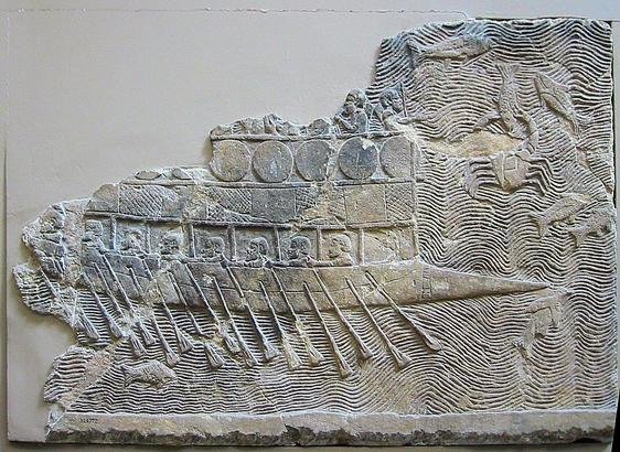 Barco de guerra fenicio en relieve asirio del palacio de Nínive. El barco tiene dos niveles. En el inferior, se ve dos hileras de remos y las cabezas de los remeros. Arriba, hay unos escudos o círculos y hay soldados, pero no se ve mucho más porque no se ha conservado. En el agua hay varios peces y un cangrejo.