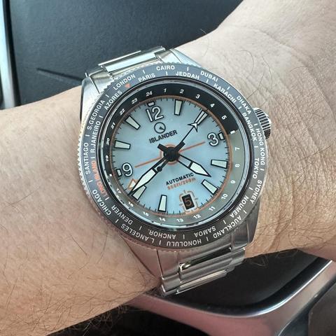 Watch with brushed stainless steel case with polished chamfer, brushed stainless steel H-link bracelet, world time bezel, pale blue dial with white numeric indices outlined in black at 12, 3 and 9, orange-on-brown date at 6:00 with date window outlined in orange, baton indices elsewhere, skeletonized black sword hands with white lume, orange GMT hand, arrow seconds hand, and 24-hour markings on the rehaut. 