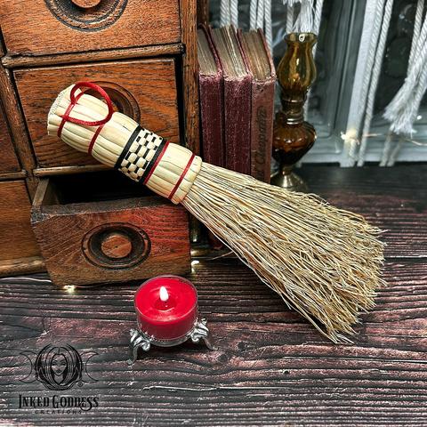An Altar Besom is propped against a wooden cabinet on a dark tabletop. It's made of undyed straw with red and black patterns on the woven handle. This is an exclusive product from Inked Goddess Creations.