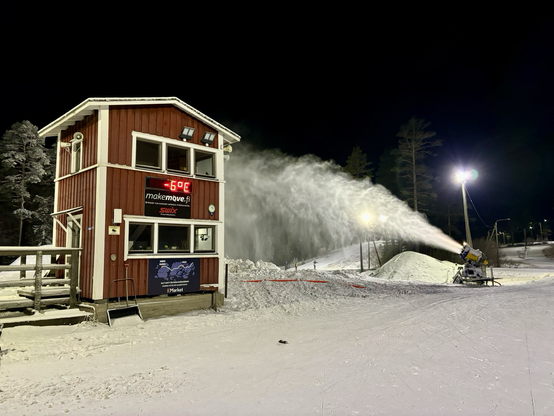 Rotes Holzhaus zeigt -6 Grad, daneben pustet eine Schneekanone