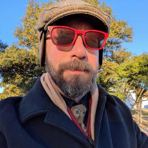 A photo of me, a 46 year old white man with a shaved head, a short cropped brown & grey beard, 

photographed from the chest up in front of some live oak trees in an office park,

wearing

a rectangle glasses with red frames, a black dress shirt, a goldtone necktie with blue sunbursts, under a deep red quarter zip sweater, khaki scarf, and black greatcoat with a light brown flat cap with earflaps.