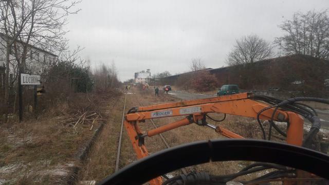 Das Bild zeigt eine Szene an einem verlassenen oder stillgelegten Gleis in der Nähe von Lüchow. Im Vordergrund steht ein orangefarbener Fiedler-Kran, der wahrscheinlich zur Beseitigung von Vegetation entlang der Gleise verwendet wird. Mehrere Personen sind im Hintergrund zu sehen, die ebenfalls an Reinigungs- oder Instandhaltungsarbeiten beteiligt scheinen. Das Umfeld wirkt vernachlässigt; die Vegetation ist üppig, und die Gebäude im Hintergrund scheinen verlassen oder in einem schlechten Zustand zu sein. Der Himmel ist grau und bedeckt, was zur insgesamt etwas melancholischen und etwas verlassenen Stimmung des Bildes beiträgt. Das Bild könnte einen Moment der Arbeit an der Revitalisierung oder dem Abriss einer stillgelegten Bahnstrecke einfangen. Der Kontrast zwischen dem modernen Kran und dem verfallenden Umfeld ist auffällig.