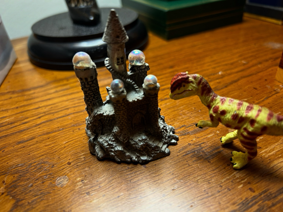 A green Dilophosaurus with brown spots looks down at a tiny pewter castle with disappointment in their eyes.