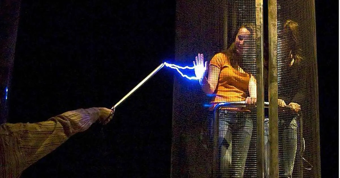 2 women inside a Faraday cage; one holds up her palm where a current arcs between the cage and a rod held by a third person.