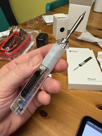 A hand holds a smart soldering tool, labeled "Pinecil," featuring a metallic tip and a transparent body with a digital display. Nearby are accessories, including wires and a box. The background shows a wooden table and a tablet.