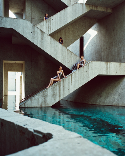 Four women in modern, avant-garde outfits pose at various levels of a dramatic concrete staircase above a pool of clear, turquoise water. The strong lines of the architectural setting and vibrant reflections underscore the bold, futuristic vibe of the scene.