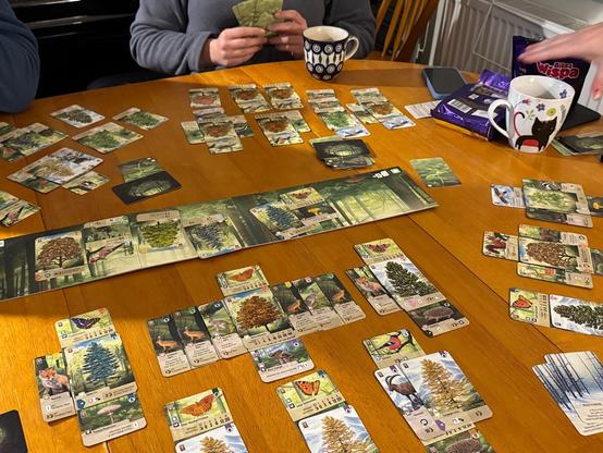 A tabletop gaming scene featuring cards spread across a wooden table, depicting trees and wildlife. Two players are holding cards, with mugs and snacks visible in the background. A colorful game board is displayed in the center.