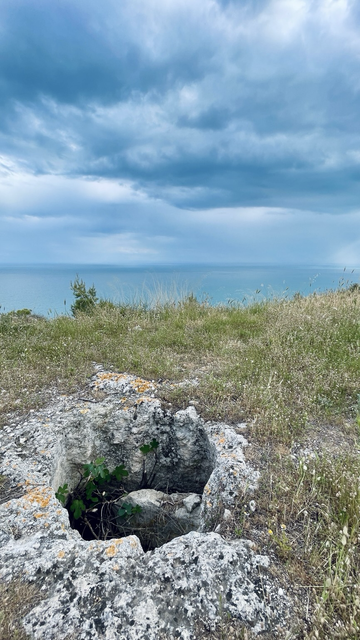 Im Vordergrund ein Grab in den Felsen geschlagen, dahinter grüne Vegetation. Im Hintergrund das Meer (Adria)