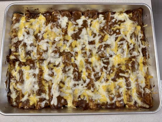 A big metal baking pan full of cheese and ground beef enchiladas, smothered in traditional chili enchilada sauce and shredded cheese. 