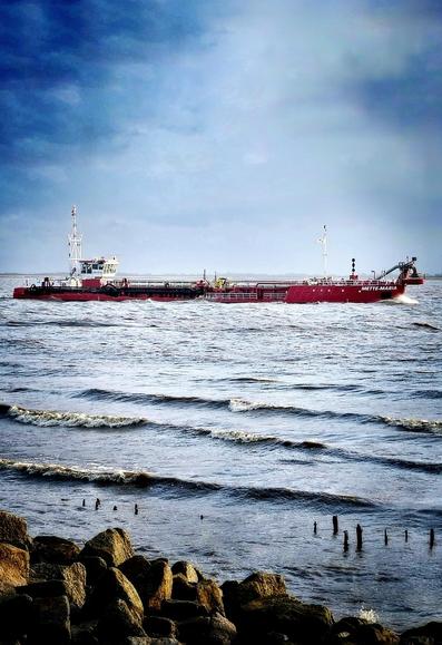 The photo shows a large red hopper dredger sailing on a wide river. The sky is cloudy with dramatic lighting, creating a sense of depth and atmosphere. In the foreground, there are rocky shores with some old wooden posts sticking out of the water. The waves are visible on the water's surface.