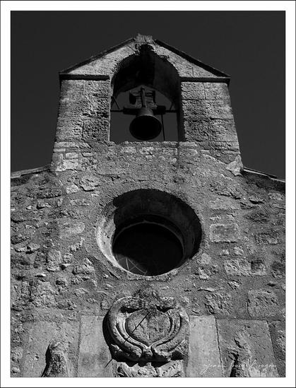 

This is a black and white image of an old stone church building. The structure features a central circular window, reminiscent of a small rose window, set into a weathered stone facade. Above the window, there's a bell housed within a simple open-air belfry, framed by a tapered stone arch. Beneath the window, there's a decorative element, possibly a carved emblem or crest. The overall appearance gives a sense of antiquity and historical architecture.