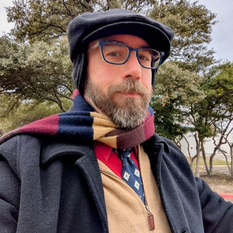 A photo of me, a 46 year old white man with a shaved head, a short cropped brown & grey beard, 

photographed from the chest up in front of some live oak trees in an office park,

wearing

a rectangle glasses with dark blue frames, a bright red dress shirt, a dark blue necktie with white arabesque flowers , under a khaki cardigan, a of navy, red, and tan, and black greatcoat with a black flat cap with earflaps