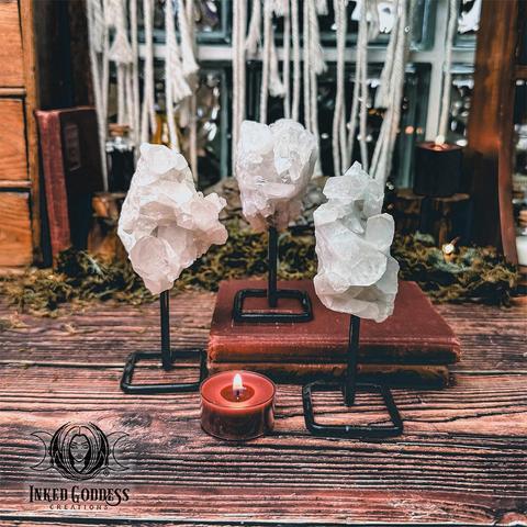 Three specimens of the "Quartz Point Cluster on Metal Stand" product are arranged around a tealight candle and old books. The metal stands are round black iron formed into a square at the base with a single stem going up to the crystals. Presented by Inked Goddess Creations.