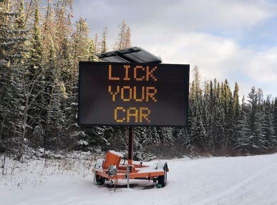An LED road sign sitting in the snow, reading "LICK YOUR CAR"