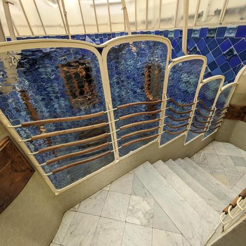 Curvy wooden handrails on steps going downward with blue tile in the background visible through the wavy glass walls. 