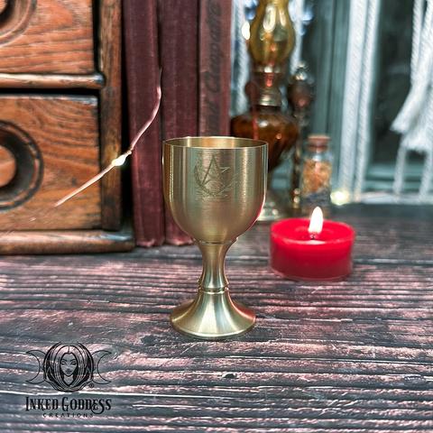 A Mini Brass Pentacle Chalice stands on a dark tabletop. It's made of shiny brass and it has a pentacle and laurel boughs engraved on the cup. This is an exclusive product from Inked Goddess Creations.