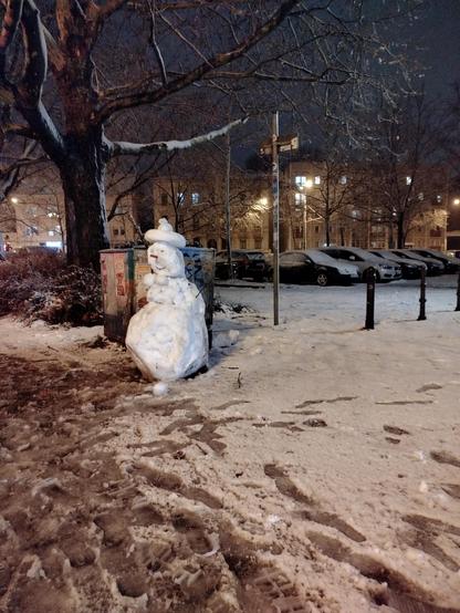An der dunklen Breiten Straße in Potsdam steht ein selbstgebauter weißstrahlender schneemann mit Hut!