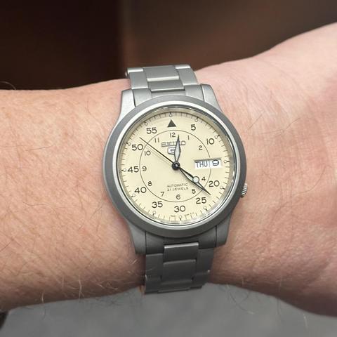Watch with a blasted stainless steel case, beige dial, hour markings in an inner circle and minute markings along the outside (Flieger type B configuration), black sword hands filled with lume, black second hand with lumed counterbalance, and 4:00 crown, on a blasted stainless steel oyster-type bracelet. 