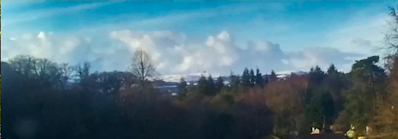 A view to the horizon, in the foreground a wood of trees with leaves of green and brown. In the background stands Hounds Tour, snow covered as the clouds roll over.