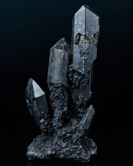 A vertical, dark quartz cluster with multiple pointed crystals protruding from a rough, rocky base, set against a black background.